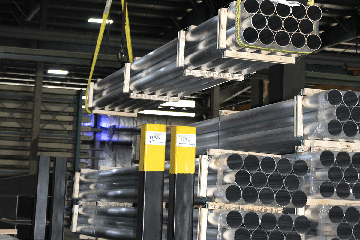marmon-keystone Marmon Keystone Bundles of black metal tube pipes are stacked on pallets in an industrial warehouse. A crane lifts one bundle while others are stored on the ground and metal shelves. Yellow safety posts are visible in the foreground.