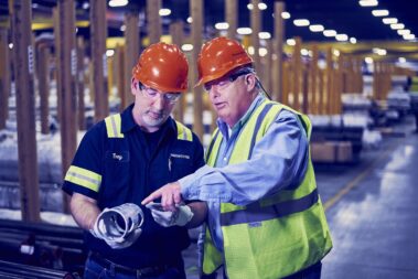 Marmon Keystone Two men wearing orange hard hats and safety glasses stand in a factory. One holds a metal tube as the other, in a safety vest, points at it, discussing or inspecting the item as part of the supplychain in a metals distributor setting.