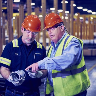 Marmon Keystone Two men wearing orange hard hats and safety glasses stand in a factory. One holds a metal tube as the other, in a safety vest, points at it, discussing or inspecting the item as part of the supplychain in a metals distributor setting.