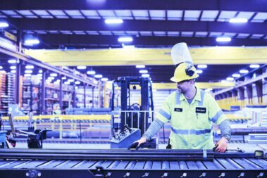 Marmon Keystone A worker in a high-visibility jacket and hard hat operates machinery on a conveyor belt in a large, well-lit industrial warehouse. Shelves of tube, pipe, and bar metals highlight the busy supplychain distributor environment.