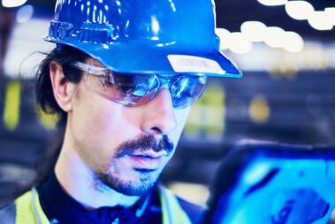 Marmon Keystone A person wearing a blue hard hat, safety glasses, and a reflective vest examines a tablet in an industrial setting, possibly managing tube and pipe supplychain operations, with blurred lights in the background.