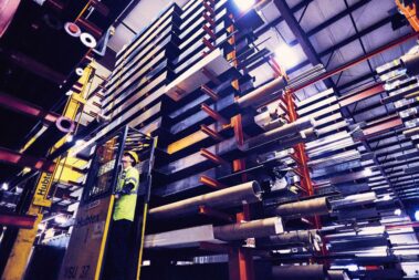 Marmon Keystone A worker in a yellow safety vest operates a forklift beside tall stacks of tube, pipe, and bar metals inside a large industrial warehouse, supporting the supplychain for a leading distributor.