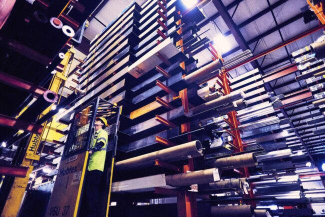 Marmon Keystone A worker in a yellow safety vest operates a forklift beside tall stacks of tube, pipe, and bar metals inside a large industrial warehouse, supporting the supplychain for a leading distributor.