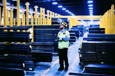 Marmon Keystone A worker wearing safety gear and a face shield stands among stacks of metal pipes and bars in a large, well-lit warehouse with yellow columns, highlighting an organized metals supply chain.