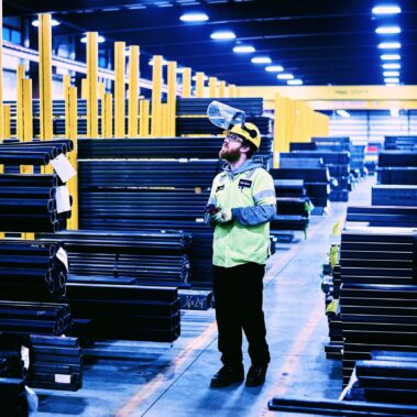 Marmon Keystone A worker wearing safety gear and a face shield stands among stacks of metal pipes and bars in a large, well-lit warehouse with yellow columns, highlighting an organized metals supply chain.