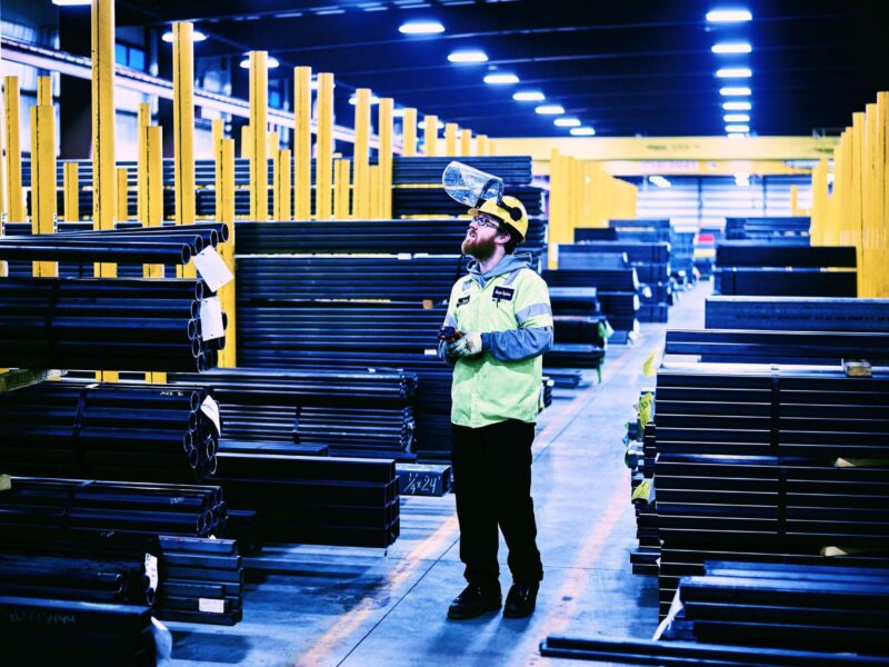 Marmon Keystone A worker wearing safety gear and a face shield stands among stacks of metal pipes and bars in a large, well-lit warehouse with yellow columns, highlighting an organized metals supply chain.