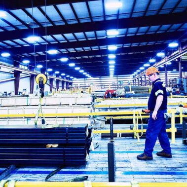 Marmon Keystone Two workers in safety gear walk near stacked metal pipes inside a brightly lit, large industrial warehouse with high ceilings, yellow safety railings, and an extensive metals supplychain.
