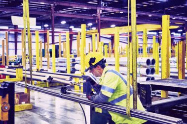 Marmon Keystone A worker wearing a yellow hard hat, safety glasses, and a high-visibility shirt operates machinery in a large, organized industrial warehouse with metal pipes and tube supplies lining the yellow safety rails.