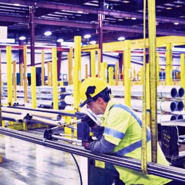 Marmon Keystone A worker wearing a yellow hard hat, safety glasses, and a high-visibility shirt operates machinery in a large, organized industrial warehouse with metal pipes and tube supplies lining the yellow safety rails.