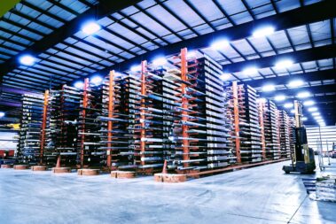 Marmon Keystone A large warehouse with tall, multi-level metal racks storing long tube pipe bars and metals. The well-lit space has a clean concrete floor and an organized supplychain layout. A forklift is visible on the right side, highlighting an efficient distributor environment.