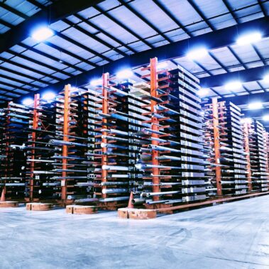 Marmon Keystone A large warehouse with tall, multi-level metal racks storing long tube pipe bars and metals. The well-lit space has a clean concrete floor and an organized supplychain layout. A forklift is visible on the right side, highlighting an efficient distributor environment.