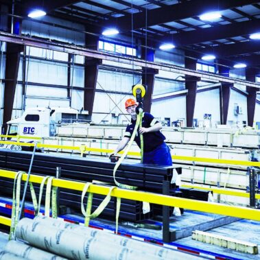 Marmon Keystone A worker in a hard hat and safety vest operates machinery to move long metal bars inside a large industrial warehouse, supporting the metals supply chain under bright overhead lights and high ceilings.