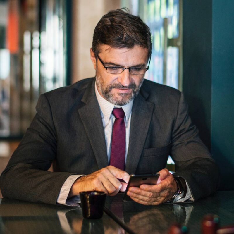 Marmon Keystone A man in a suit and tie sits indoors at a table, checking his smartphone. A small cup rests in front of him. He has glasses, short hair, and a beard—perhaps a distributor reviewing supplychain updates for metals or tube pipe bar orders.