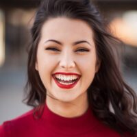 Marmon Keystone A young woman with long dark hair and bright red lipstick is smiling widely at the camera. She is wearing a red top and is photographed outdoors, near a metals distributor with a blurred background.