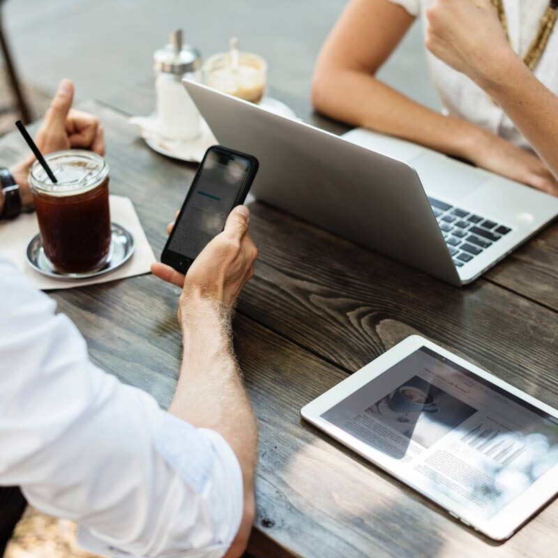 Marmon Keystone Two people sit at a wooden table outdoors with a laptop, tablet, smartphone, and iced coffee. One person types on the laptop while the other holds a phone. The tablet displays a metals supplychain distributor webpage.
