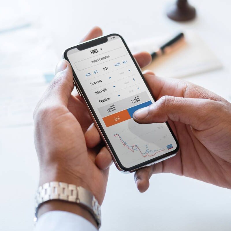 Marmon Keystone A person holds a smartphone displaying a financial trading app with stock prices, a graph, and buy and sell options—tracking metals or tube pipe bar trends. The person’s finger is about to tap "Sell." A keyboard and papers are in the background.