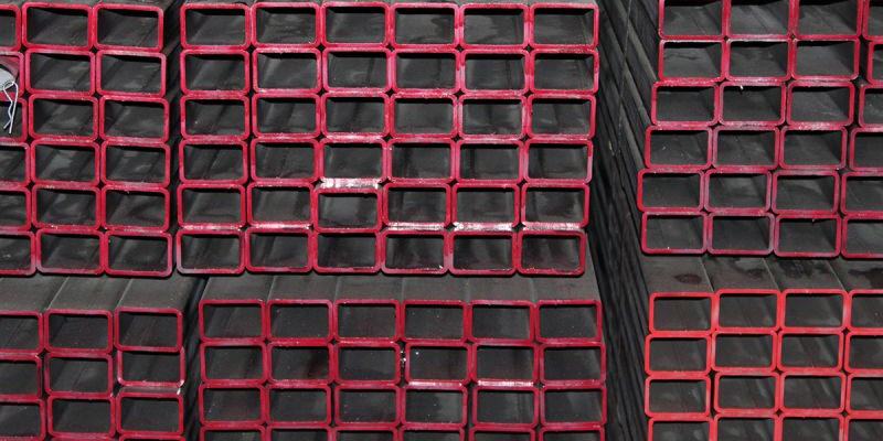 post-1-7 Marmon Keystone Stacks of rectangular hollow structural steel sections with red-painted edges are arranged in rows. The text above reads "Marmon/Keystone" and below reads "IN STOCK HSS metals distributor".