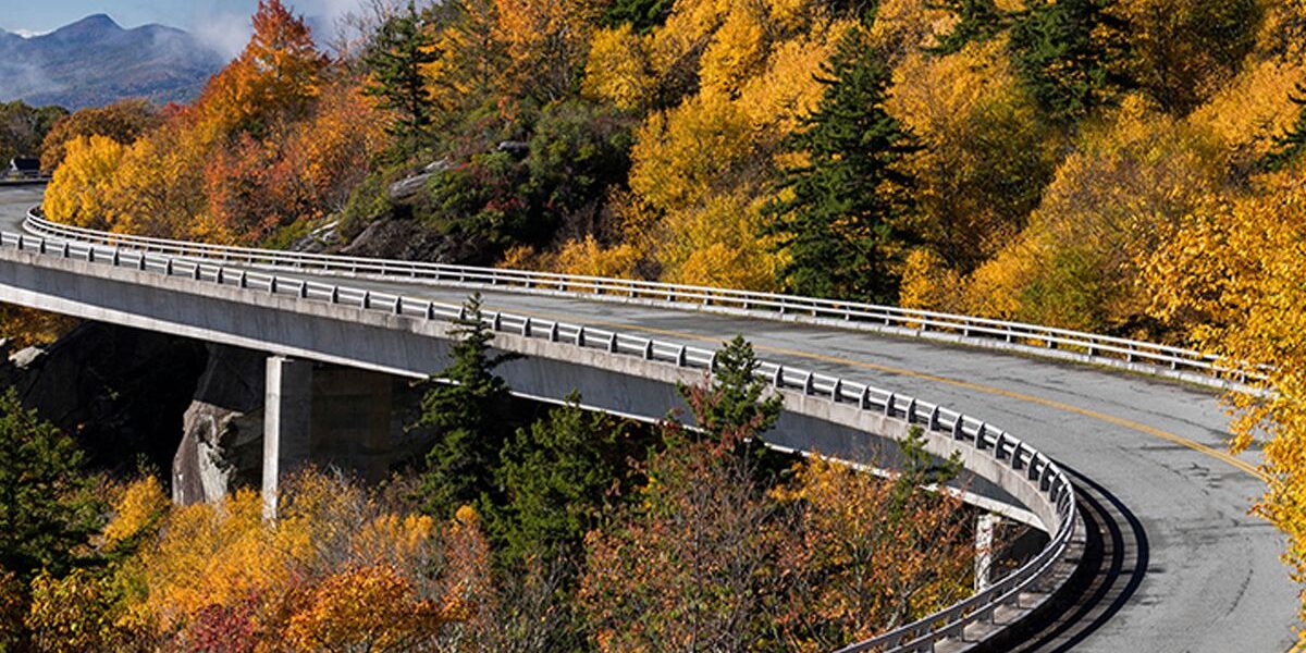 Marmon Keystone A curved highway bridge winds through a forest of autumn trees with vibrant yellow and orange foliage. Text promotes Marmon/Keystone aluminum pipe and tubing, stating Extruded Structural B429 is in stock and ready to ship.