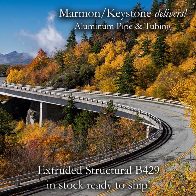 Marmon Keystone A curved highway bridge winds through a forest of autumn trees with vibrant yellow and orange foliage. Text promotes Marmon/Keystone aluminum pipe and tubing, stating Extruded Structural B429 is in stock and ready to ship.